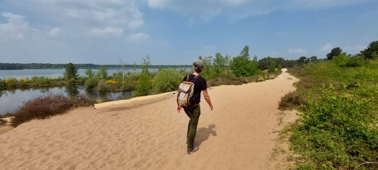 Een persoon met een rugzak loopt alleen over een zandpad naast een rustig meer, omringd door groene struiken en bomen onder een gedeeltelijk bewolkte hemel.