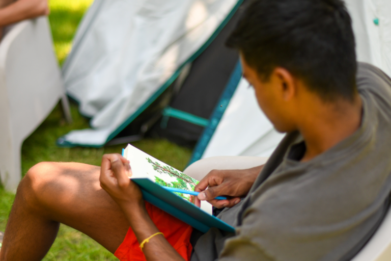 Persoon tekent in een boek, zittend in een campingstoel bij een tent op een zonnige dag.