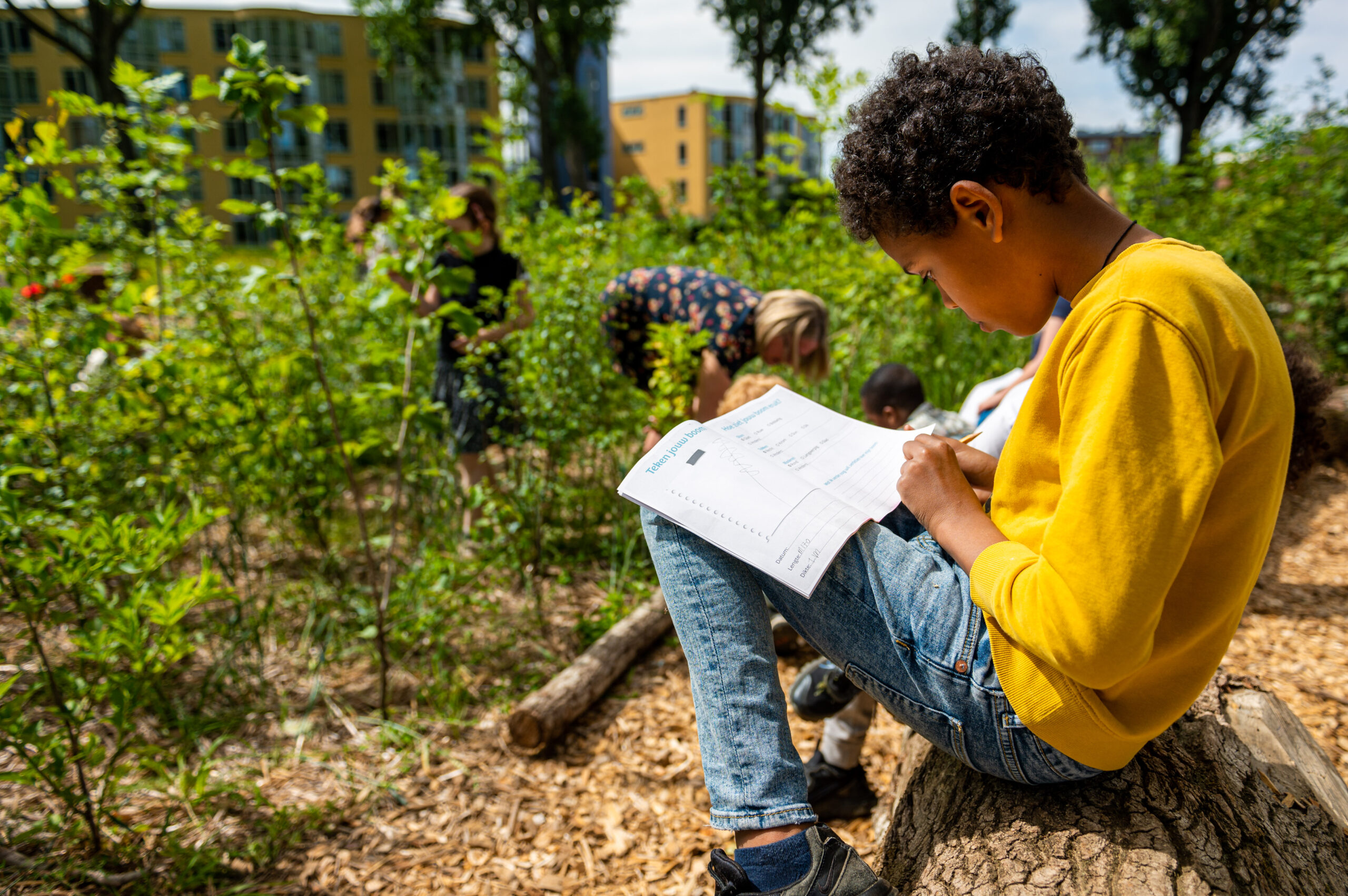 Kind leest en schrijft buiten op een boomstam, omringd door groen en andere kinderen.