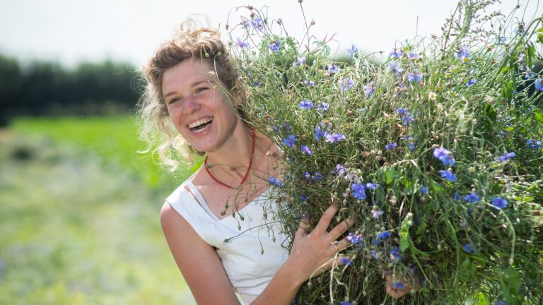 Een glimlachende vrouw in een witte jurk houdt een grote bos wilde bloemen met paarse bloesems vast in een helder, zonovergoten veld, met groen gras en bomen wazig op de achtergrond.