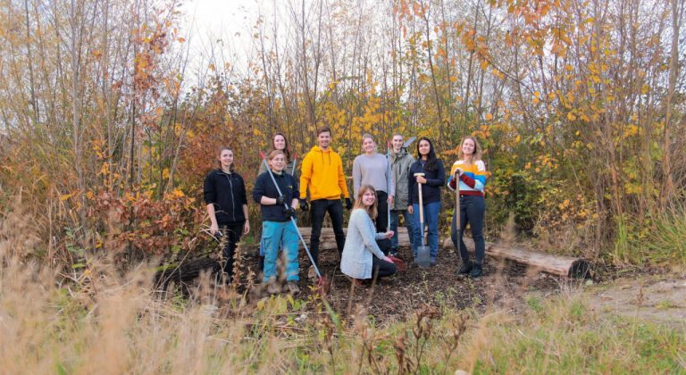 Een groep van elf mensen poseert buiten in een natuurlijke, herfstachtige omgeving met hoge grassen en kleurrijk gebladerte. De meesten staan, terwijl één persoon vooraan knielt en enkelen tuingereedschap vasthouden.