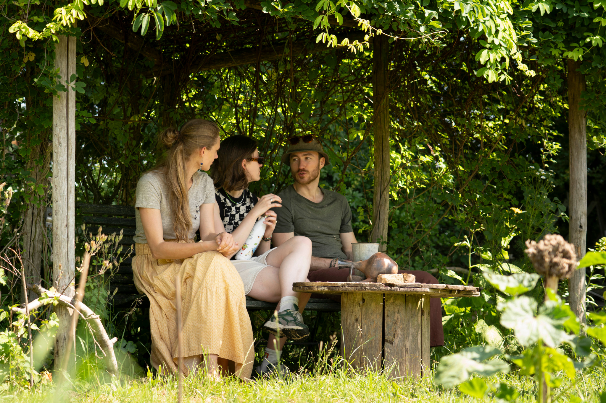 Drie personen zitten in een met groen overdekte tuinprieel en praten met elkaar.