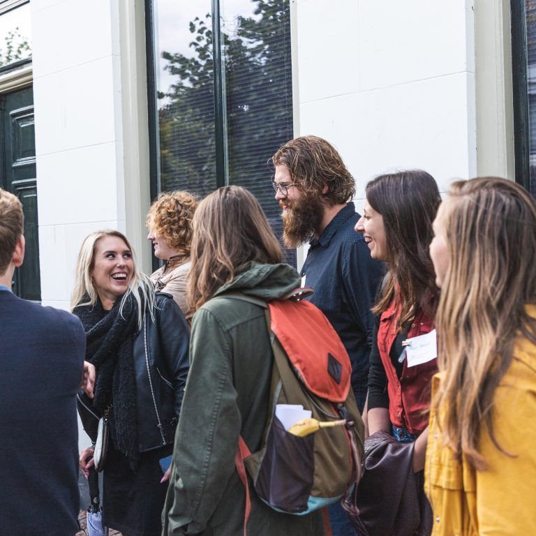 Een groep mensen lacht en praat buiten bij een gebouw met glazen deuren.