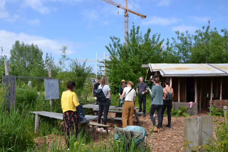 Groep mensen in een gemeenschappelijke tuin met houten schuur en bouwkraan op de achtergrond.