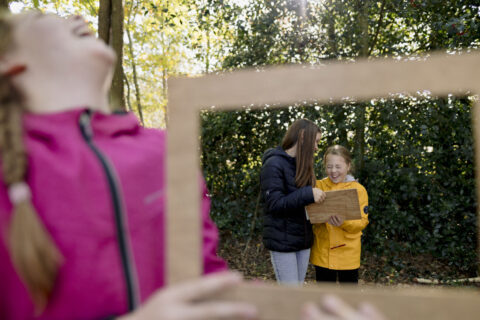 Kinderen lachen buiten met een houten bord, omringd door bomen en zonlicht.