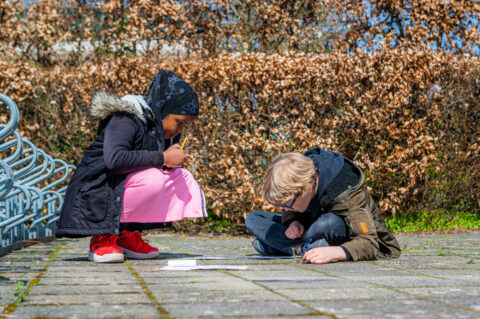 Twee kinderen tekenen buiten op de stoeptegels met potlood en papier onder een zonnige hemel.