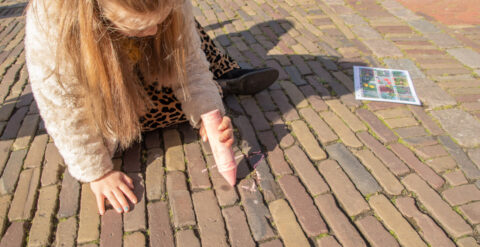 Kind tekent met roze krijt op straatstenen; naast haar ligt een blad met afbeeldingen.