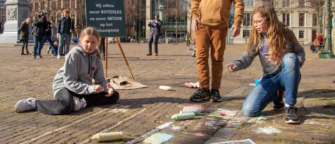 Kinderen tekenen op stoep met krijt; bord met tekst over buitenspelen op achtergrond.