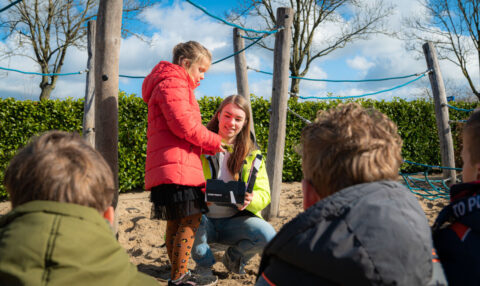 Een vrouw helpt een kind op een speeltuin bij zonnig weer, terwijl andere kinderen toekijken.