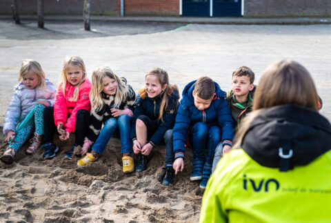Kinderen zitten in een rij op een zandvlakte, begeleid door iemand in een groene jas met "IVN".