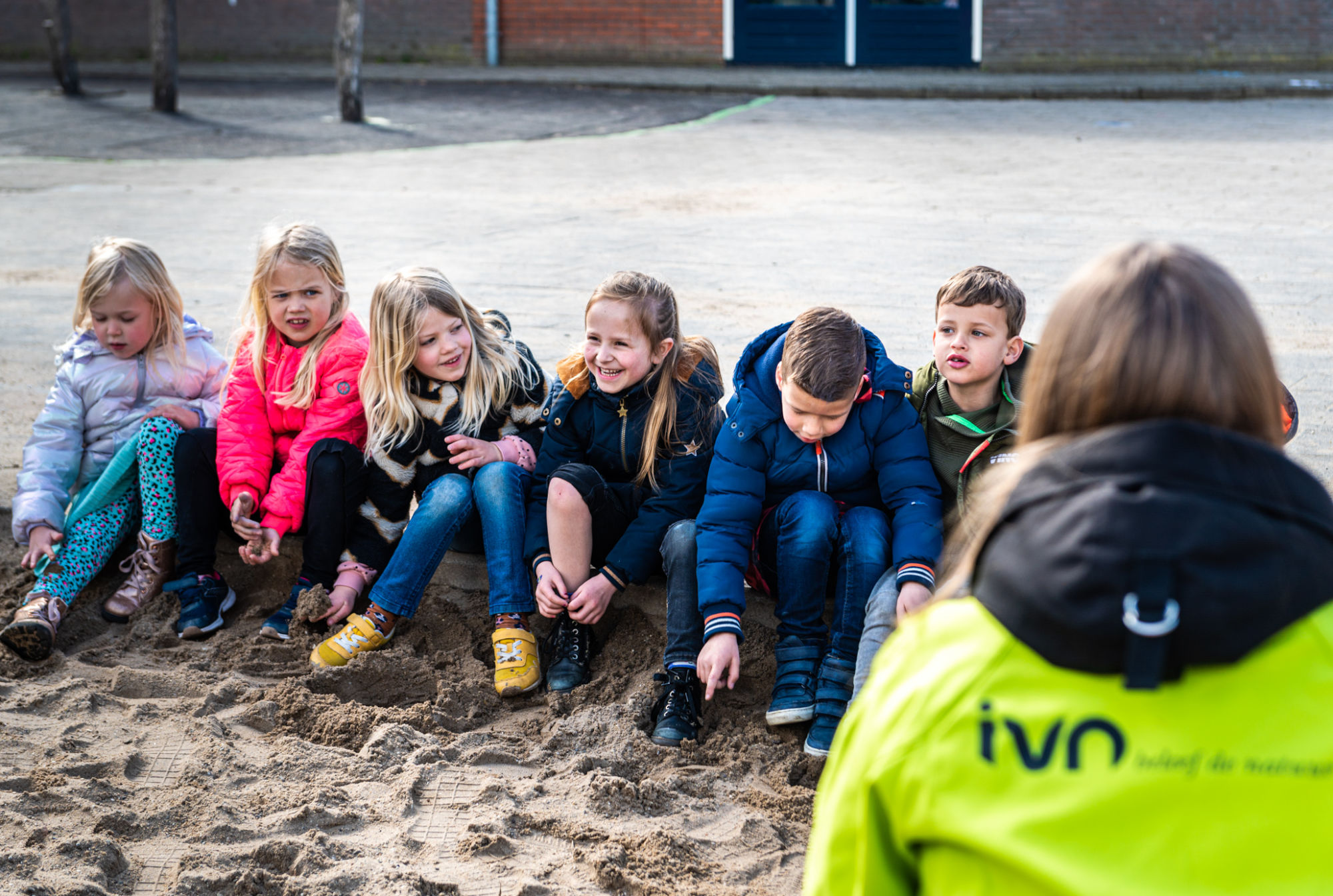 Kinderen zitten in een rij op een zandvlakte, begeleid door iemand in een groene jas met "IVN".