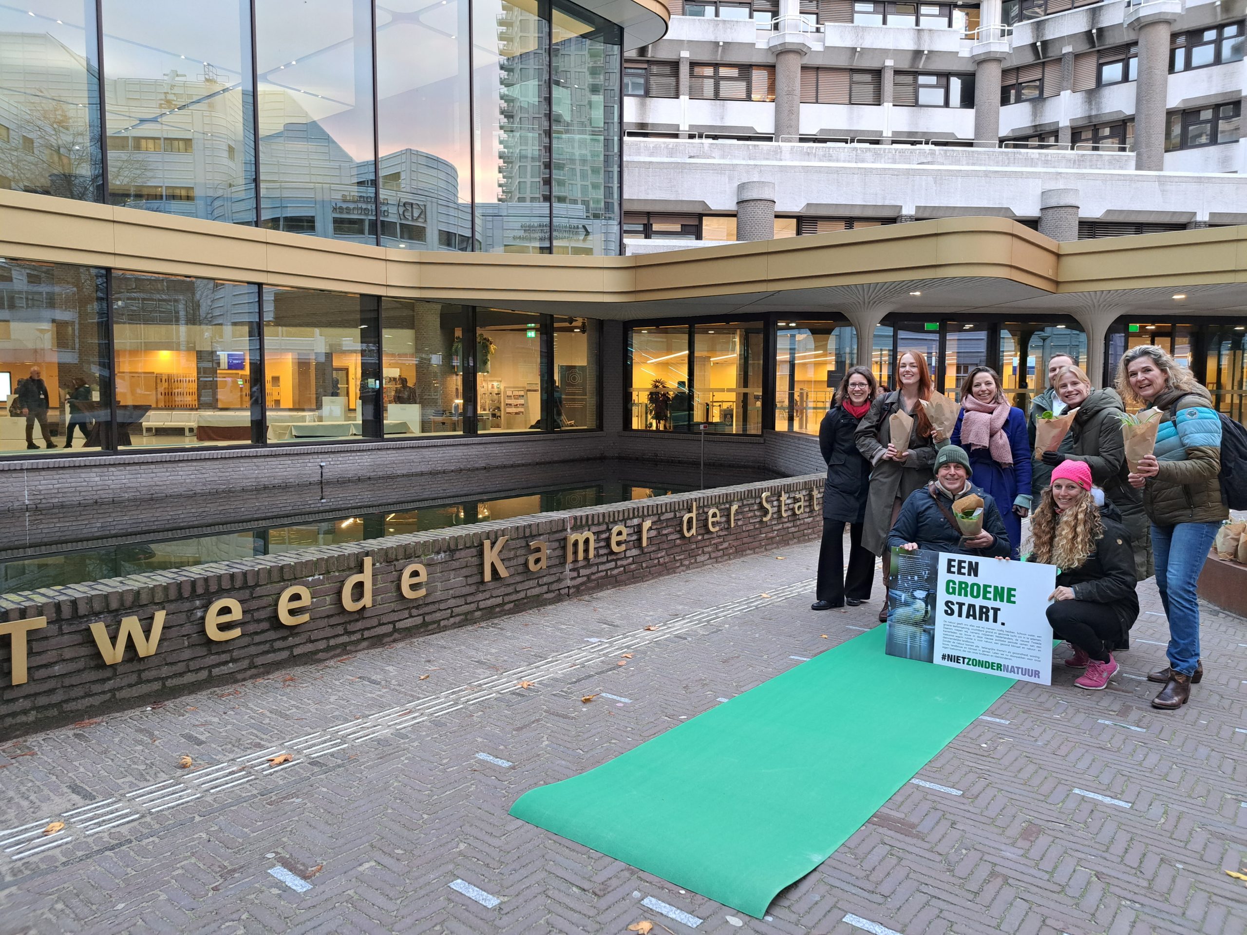 Groep mensen met een bord "Een groene start" voor het gebouw van de Tweede Kamer, naast een groene loper.
