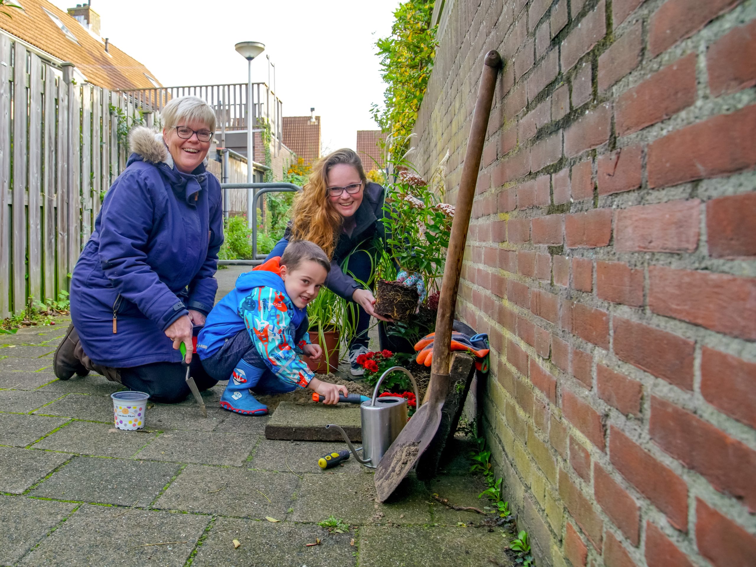 Een vrouw, kind en jongere planten bloemen langs een bakstenen muur in een steegje.