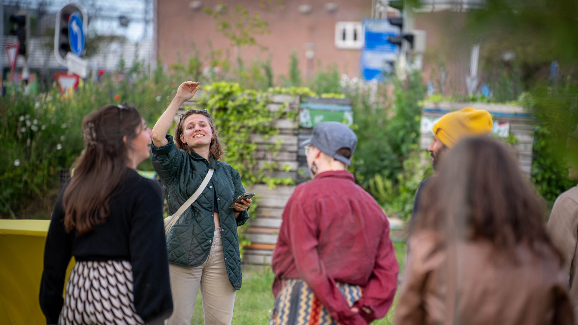 Groep mensen in gesprek in een groene tuin, vrouw met mobiel glimlacht en gebaart.