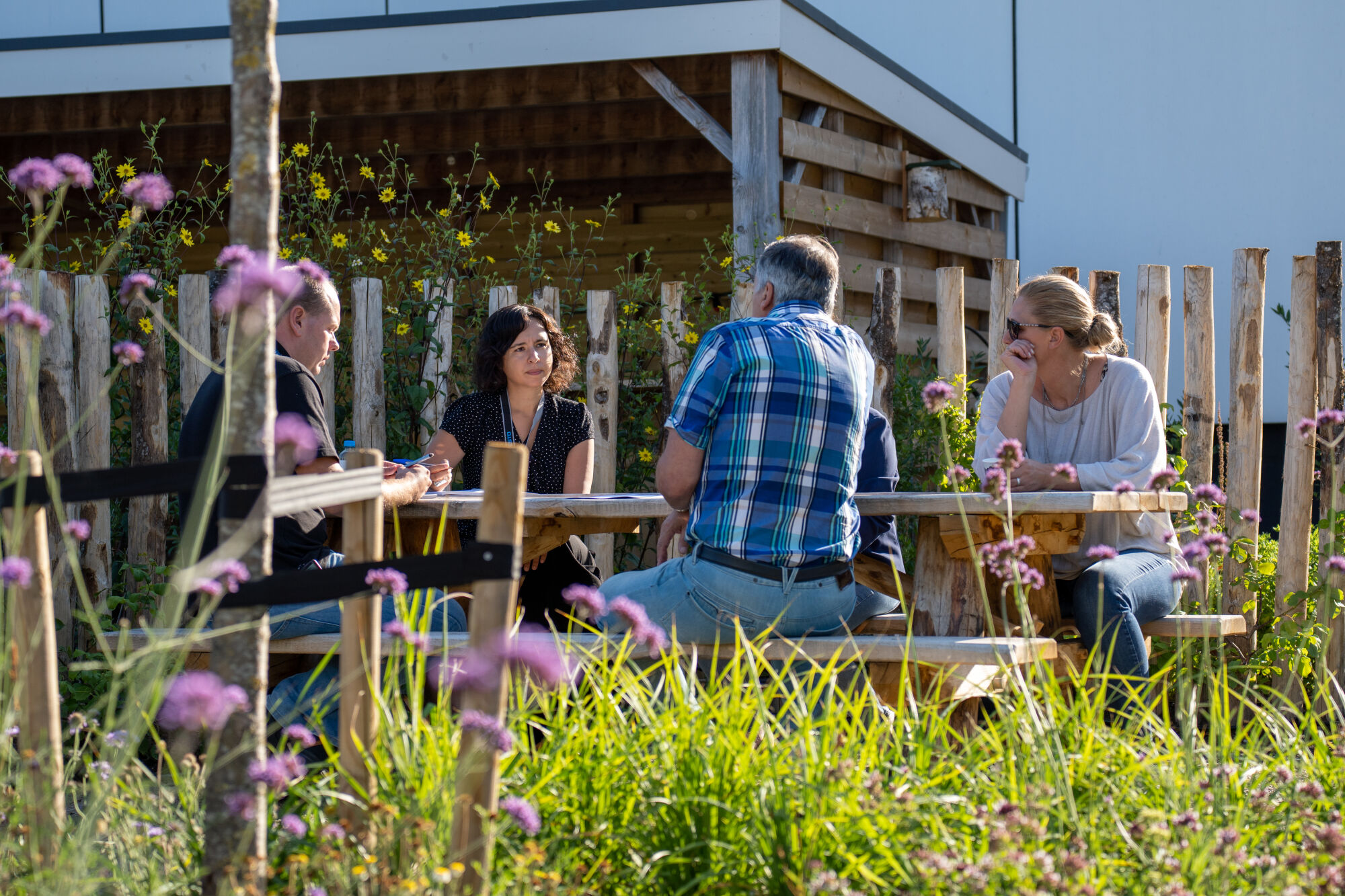 Vier mensen praten aan een houten picknicktafel in een tuin vol bloemen.