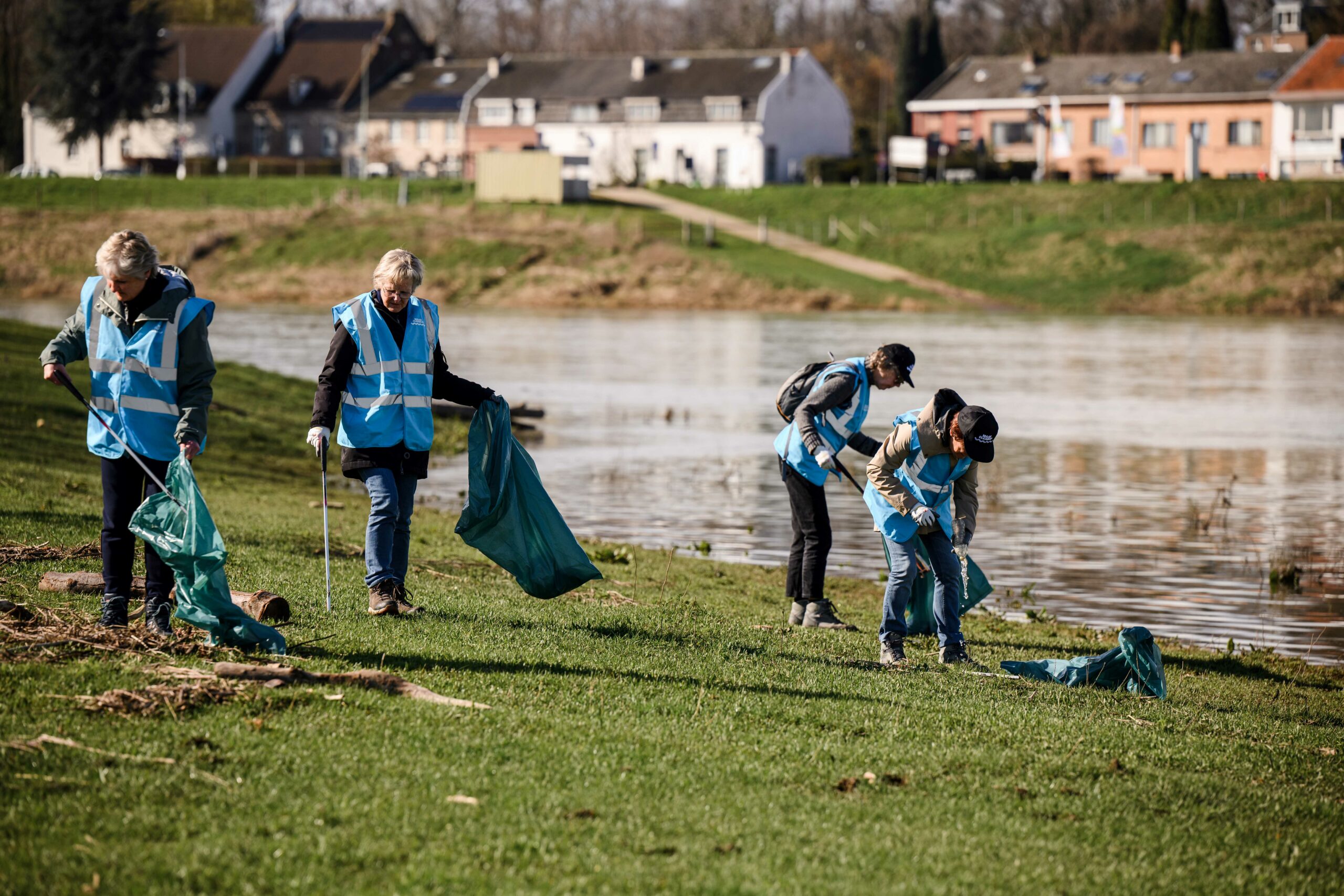 Vrijwilligers in blauwe hesjes verzamelen afval langs de waterkant in een groene omgeving.