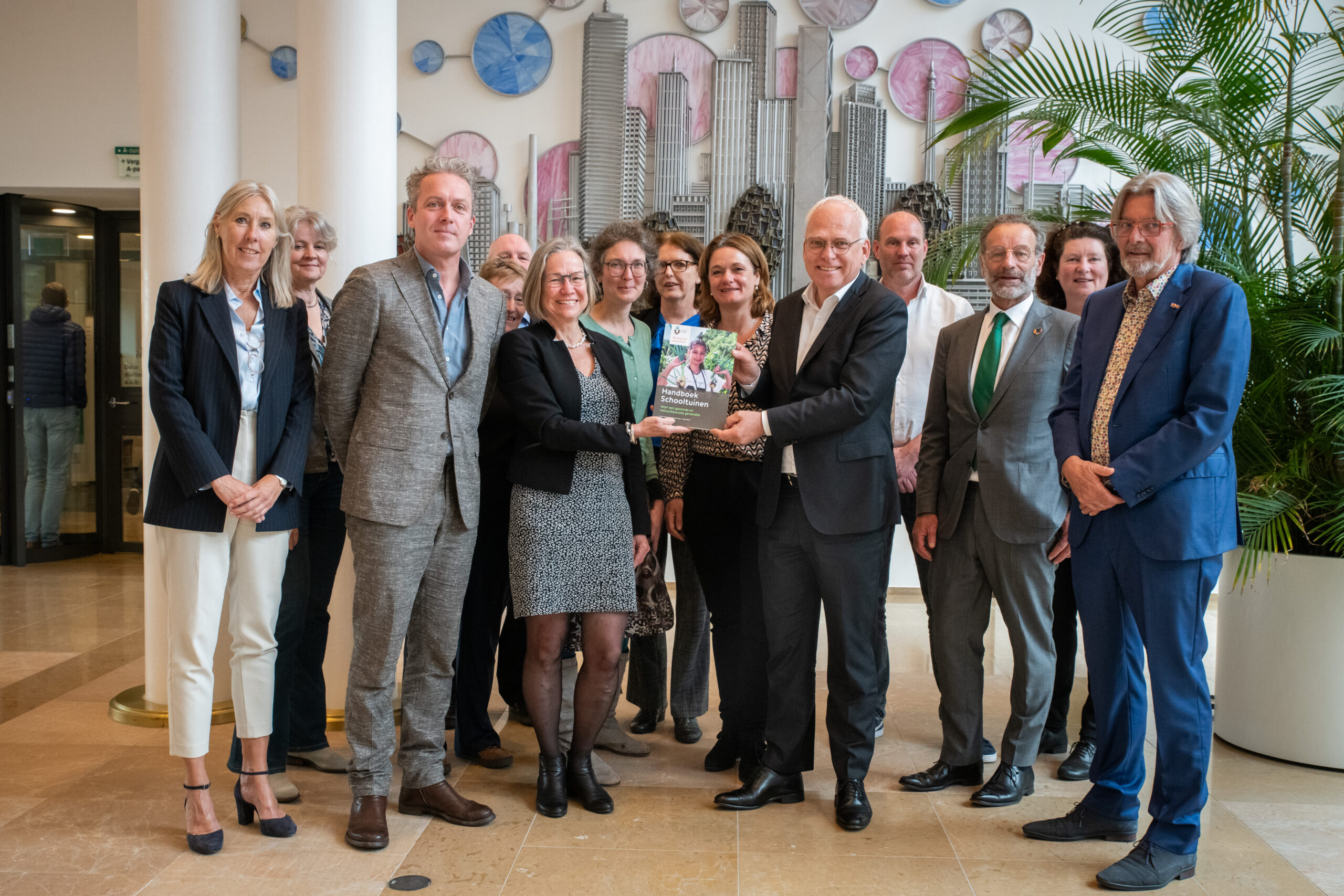 Groep mensen poseert met een boek in een gebouw, moderne kunst aan de muur.