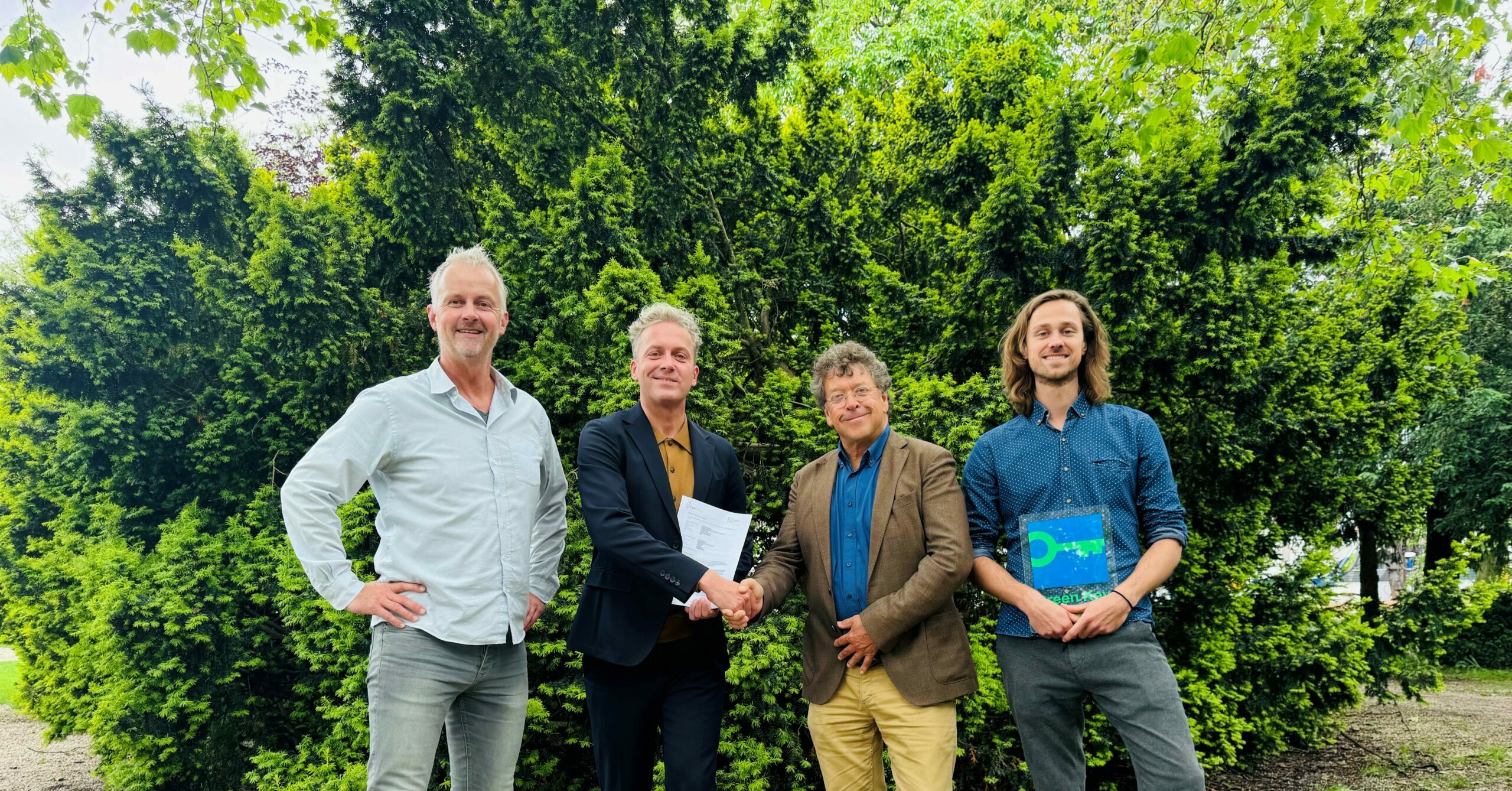 Vier mannen poseren buiten voor groene struiken, met een handdruk tussen twee van hen.