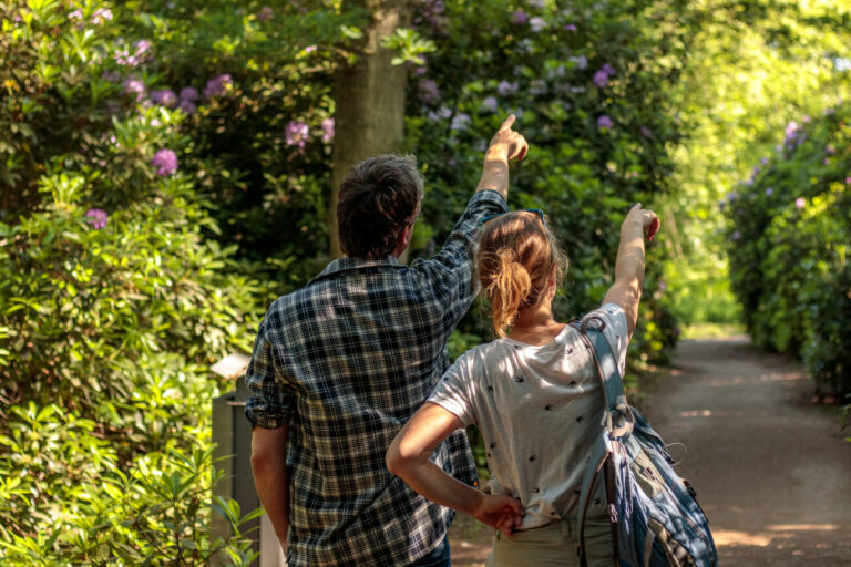 Twee mensen wijzen naar iets in een groene tuin met bloeiende struiken.