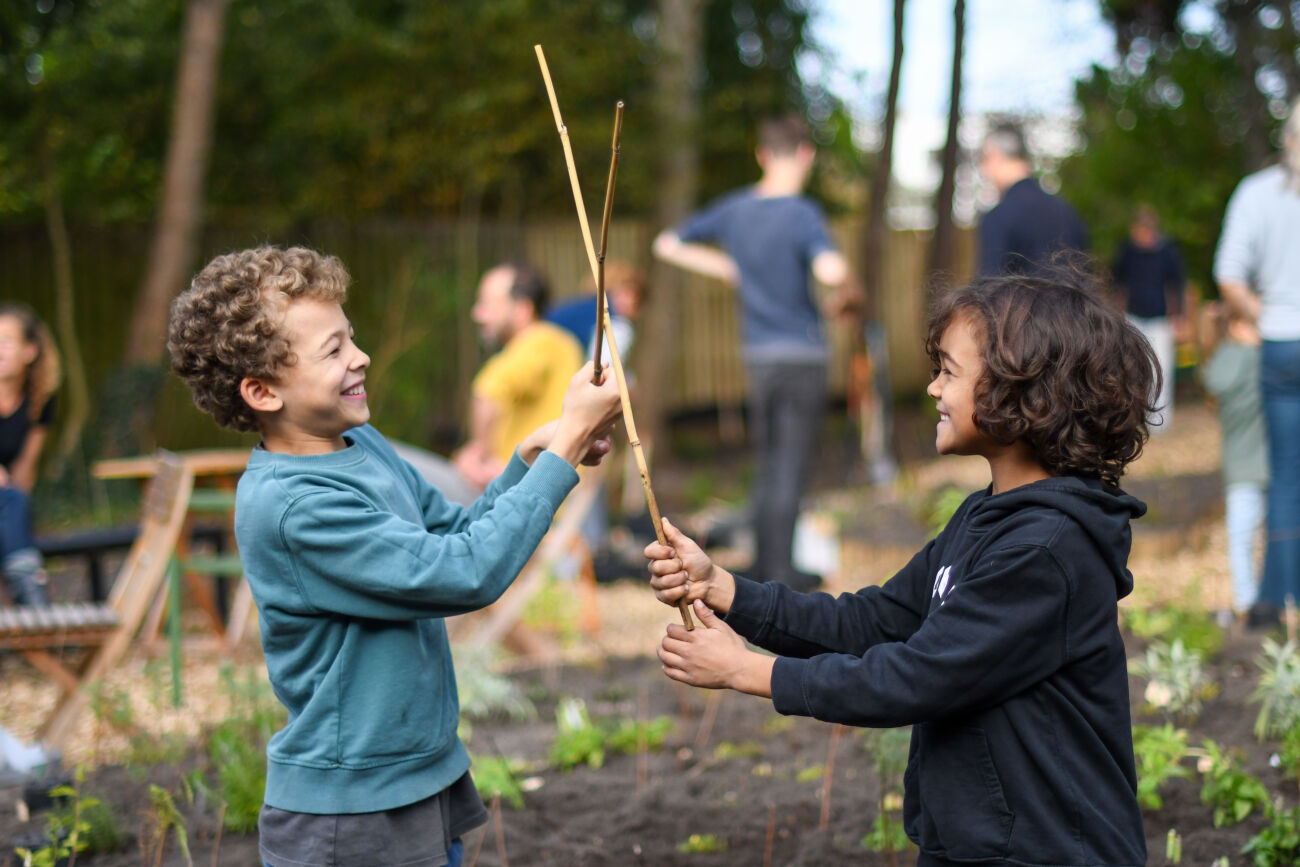 Twee kinderen spelen lachend met stokken in een tuin, omringd door volwassenen.