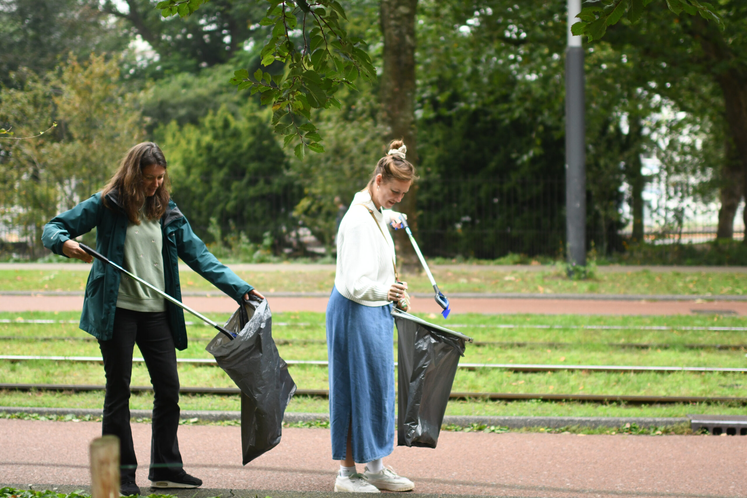 Twee vrouwen rapen afval op langs een park met grijpstokken en vuilniszakken.