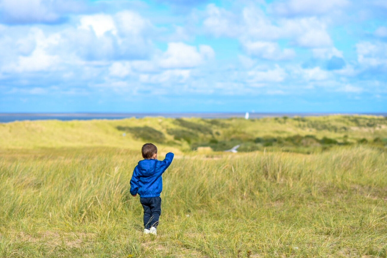 Een kind in blauwe jas loopt over grasduinen onder een bewolkte hemel.