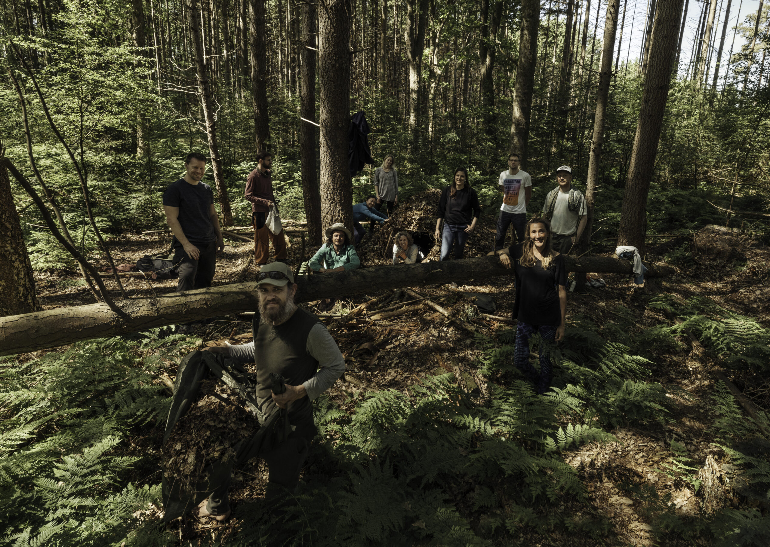 Groep mensen in een bos, verzameld rond een omgevallen boomstam en een natuurlijke hut.