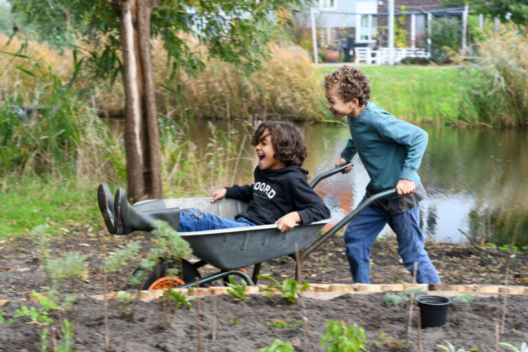 Twee lachende kinderen spelen met een kruiwagen in een tuin bij een vijver.