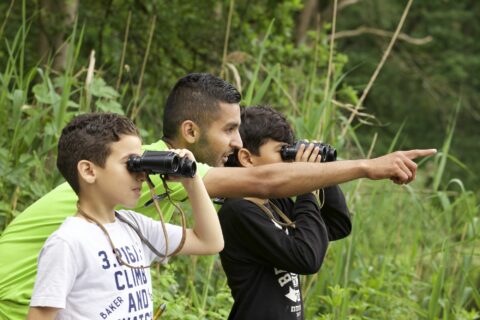 Twee kinderen en een man kijken met verrekijkers naar iets in een groene, bosrijke omgeving.
