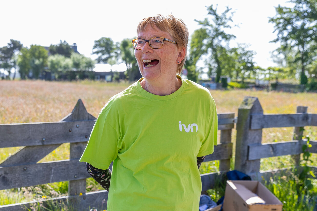 Persoon in groen shirt met IVN-logo lacht buiten bij hek en grasveld.