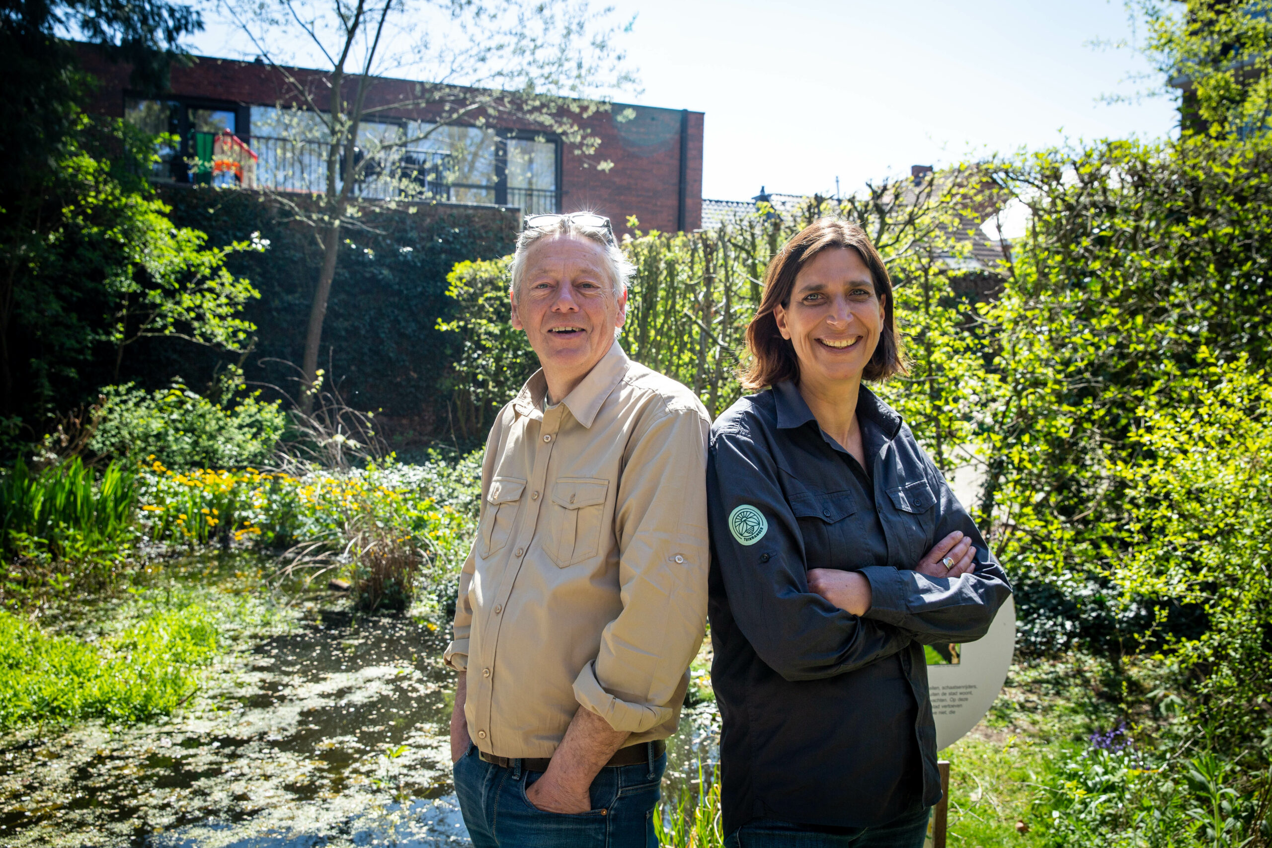 Twee lachende mensen poseren voor een vijver in een groene tuin op een zonnige dag.