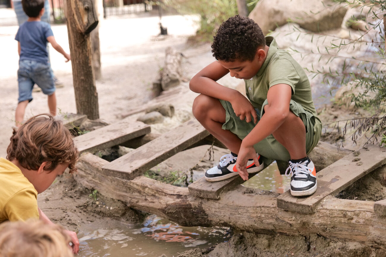 Kinderen spelen bij een modderige plas water, een jongen hurkt op een houten balk.