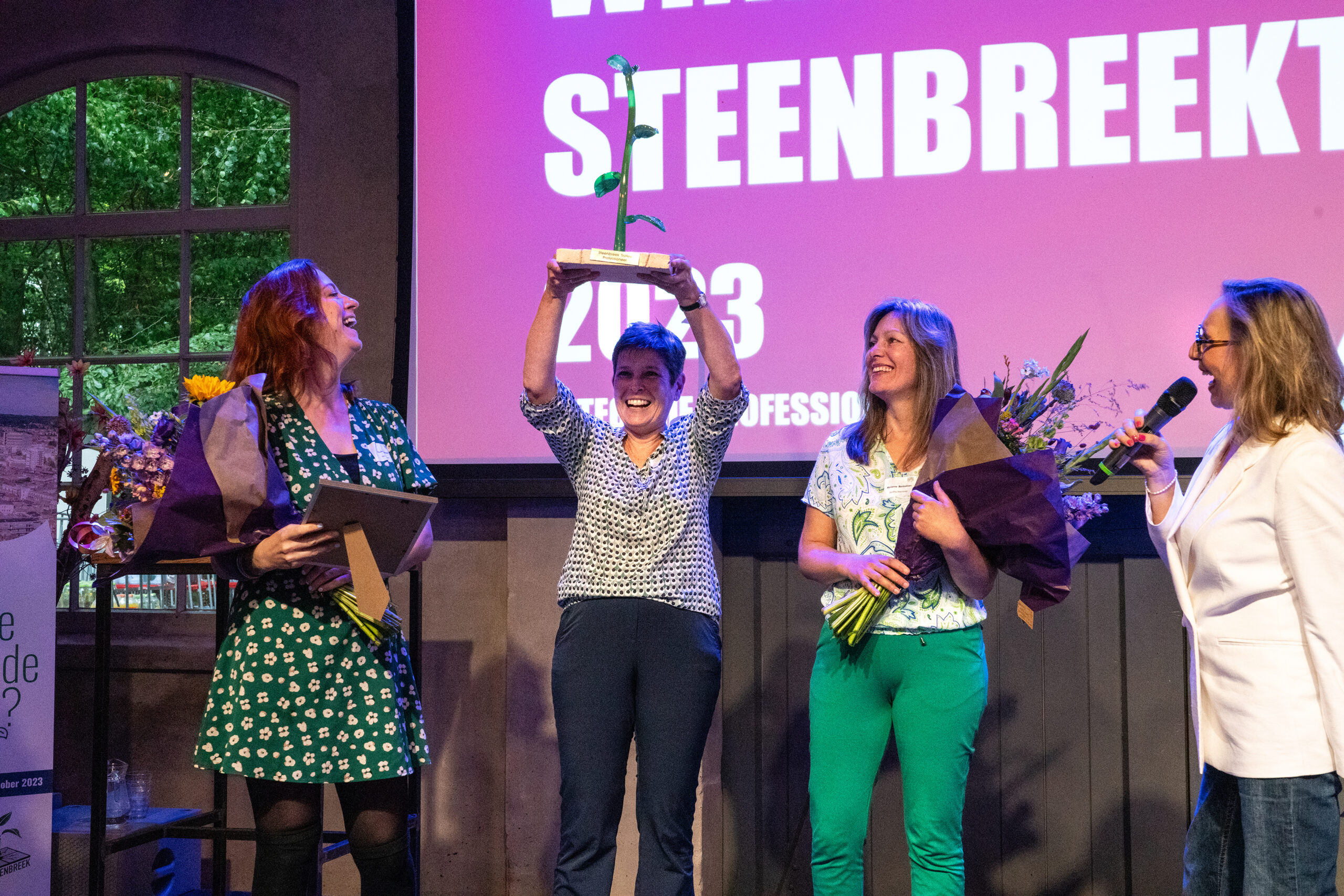 Groep vrouwen viert prijsuitreiking; winnaar houdt trofee omhoog.