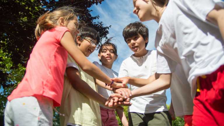 Vijf kinderen staan buiten in een cirkel, lachend en hun handen op elkaar gestapeld.