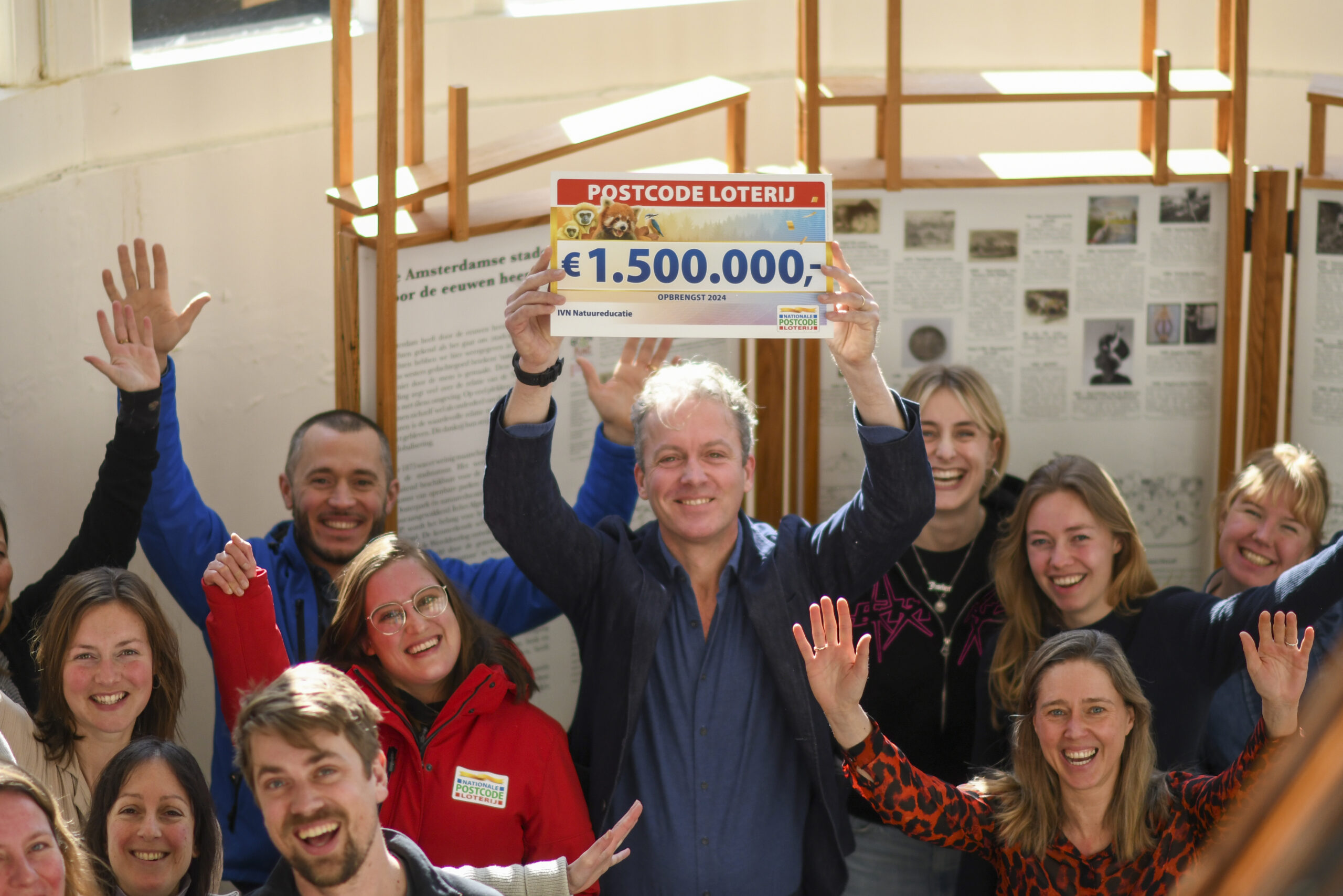 Groep mensen viert een Postcode Loterij winst van €1.500.000, met een blije man die een bord vasthoudt.