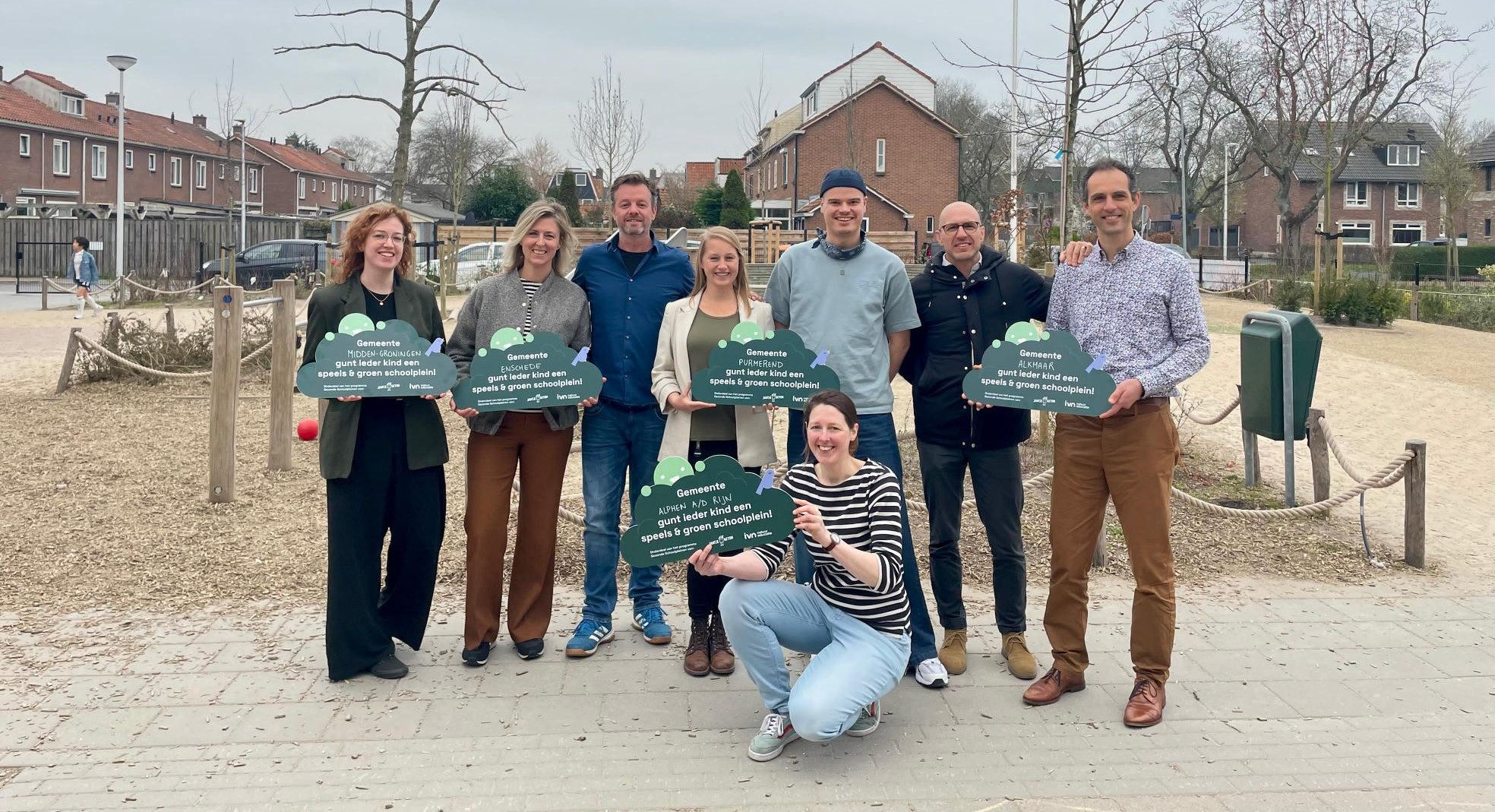 Groep mensen buiten met borden over groene, speelse schoolpleinen, omgeven door huizen en bomen.