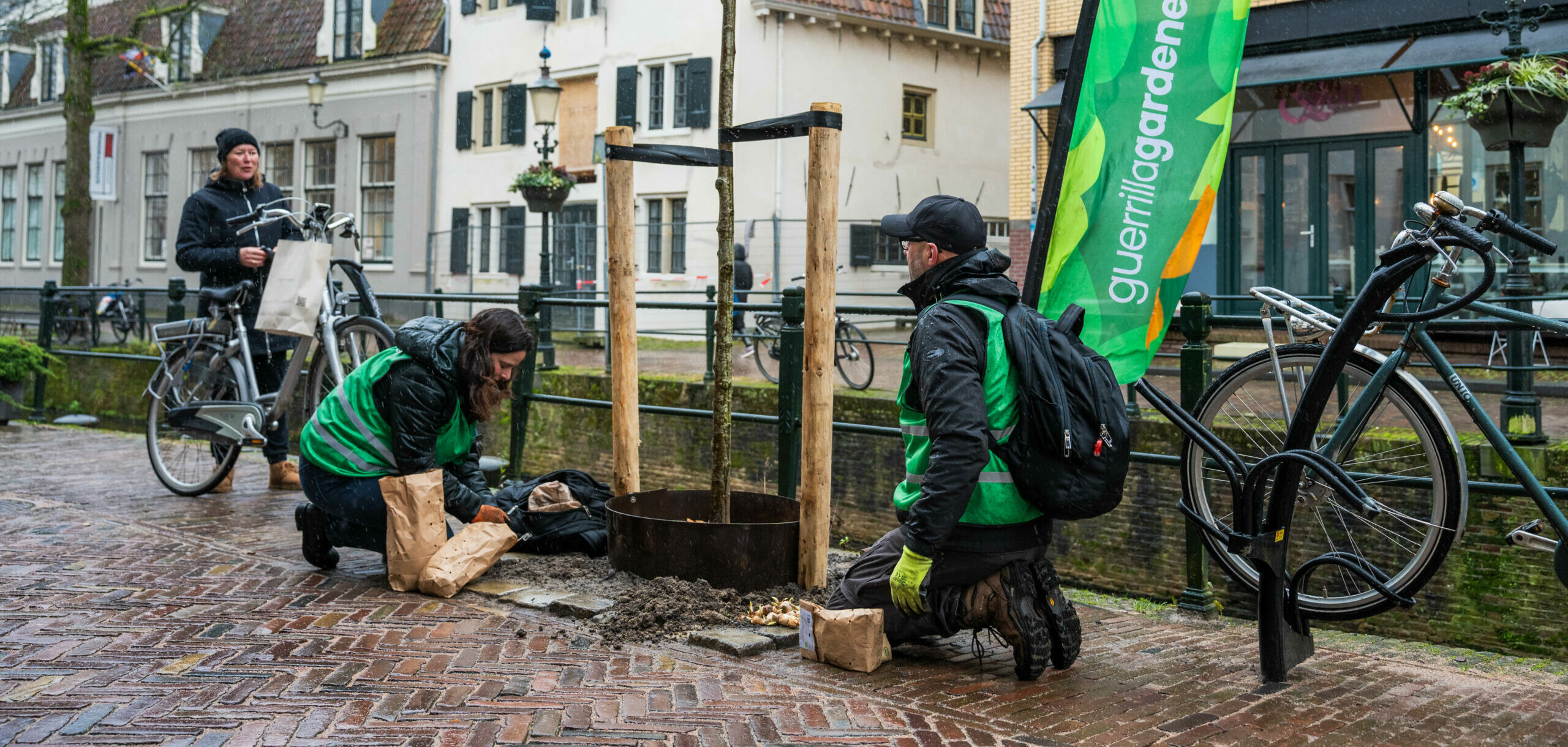 Mensen planten een boom langs de gracht met een "guerrillagardeners" vlag op de achtergrond.