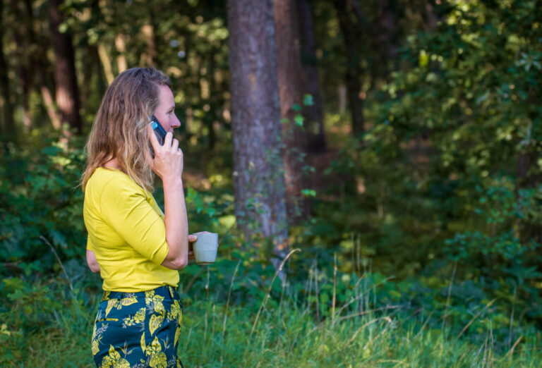 Vrouw in gele blouse belt in bos, met kopje in hand, omringd door groen.