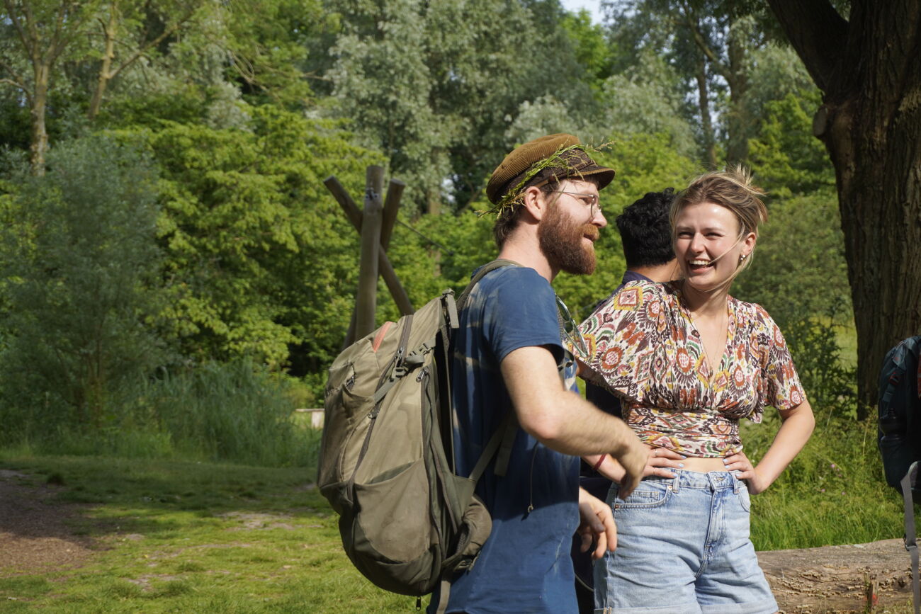 Twee mensen lachen in een groene parkachtige omgeving, man met rugzak en vrouw met kleurrijke blouse.