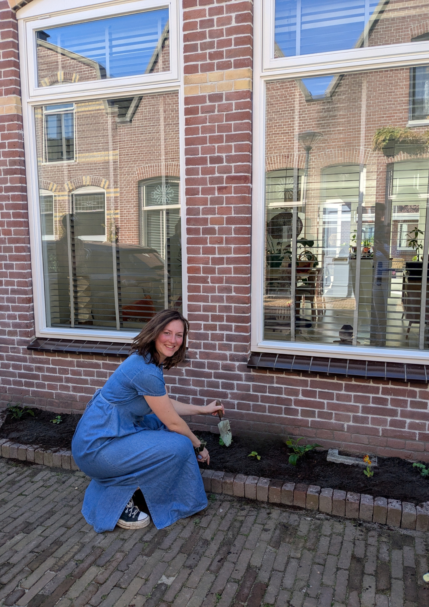 Vrouw in blauwe jurk tuiniert voor bakstenen huis met grote ramen.