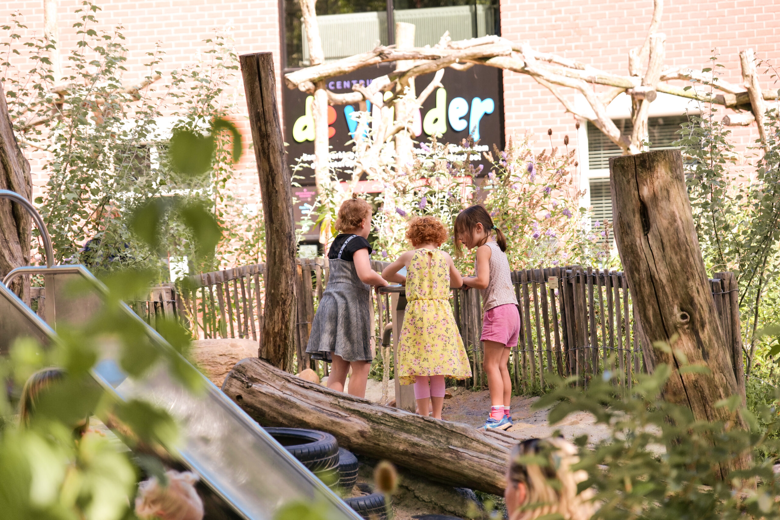Drie kinderen spelen buiten op een natuurspeelplaats met groen en houten structuren.