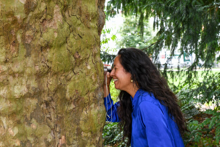 Vrouw in blauwe jas kijkt door een camera naar een boom in een groene omgeving.