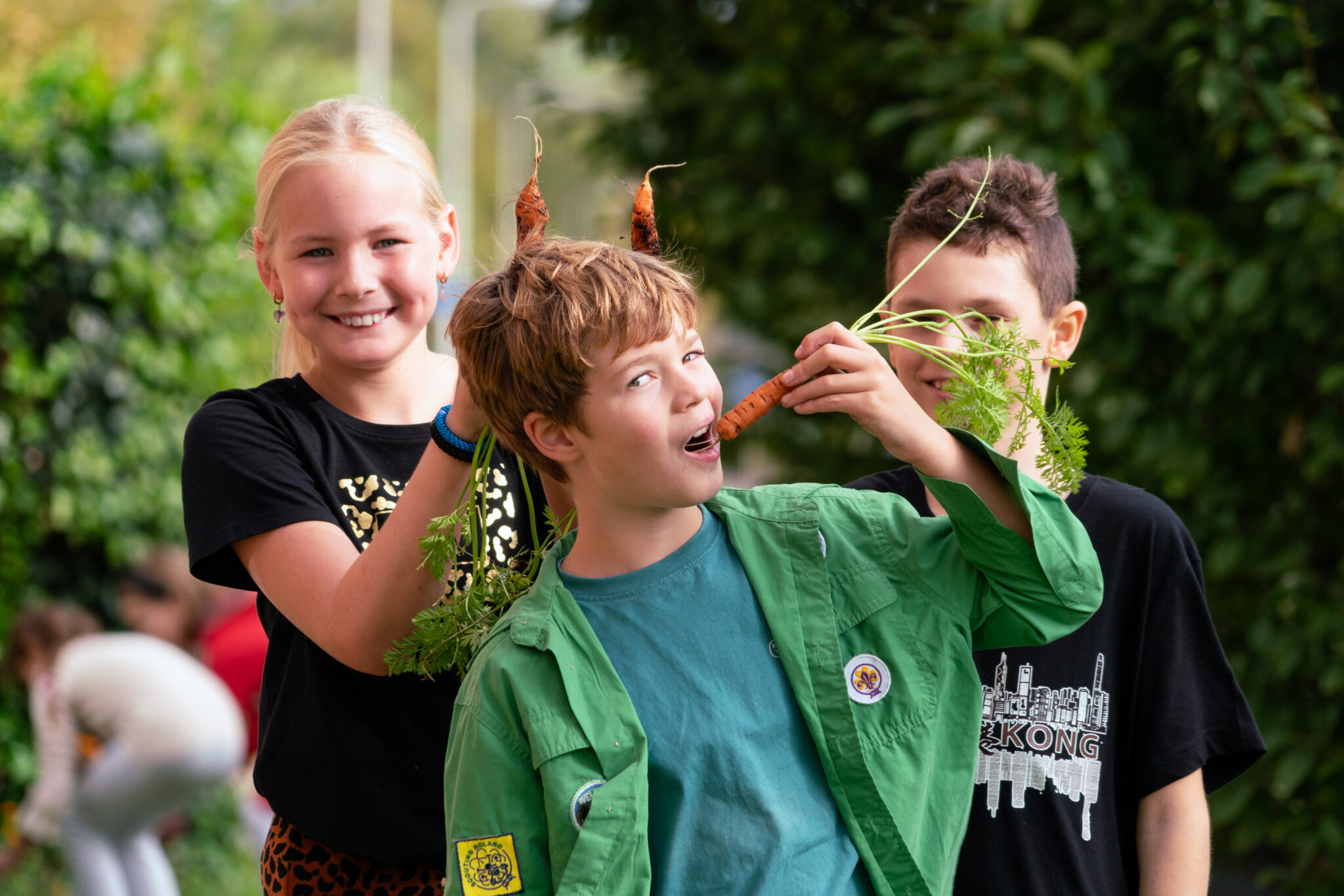 Drie kinderen spelen met wortelen in een tuin en maken grappige hoorns op hun hoofd.