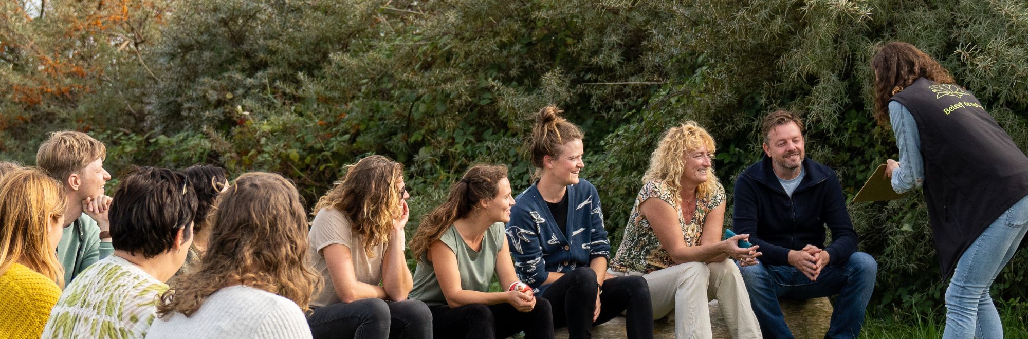 Groep mensen zit buiten in een kring, iemand staat met een clipboard tegenover hen.