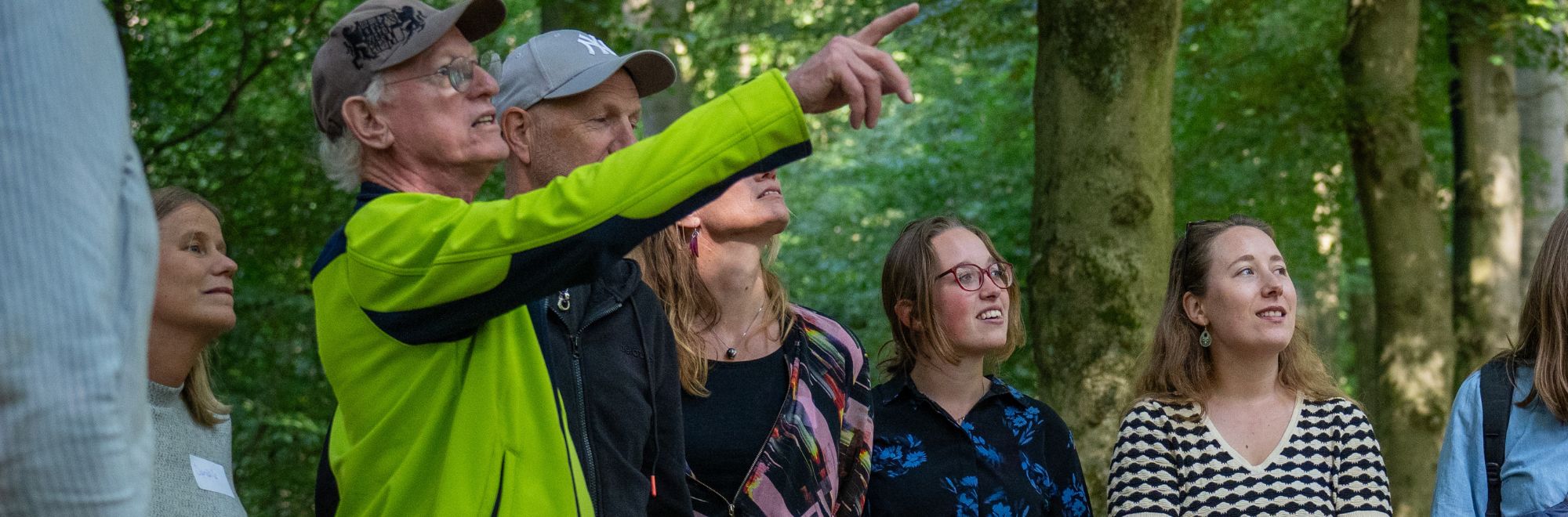 Groep mensen kijkt omhoog in een bosrijke omgeving, één persoon wijst iets aan.