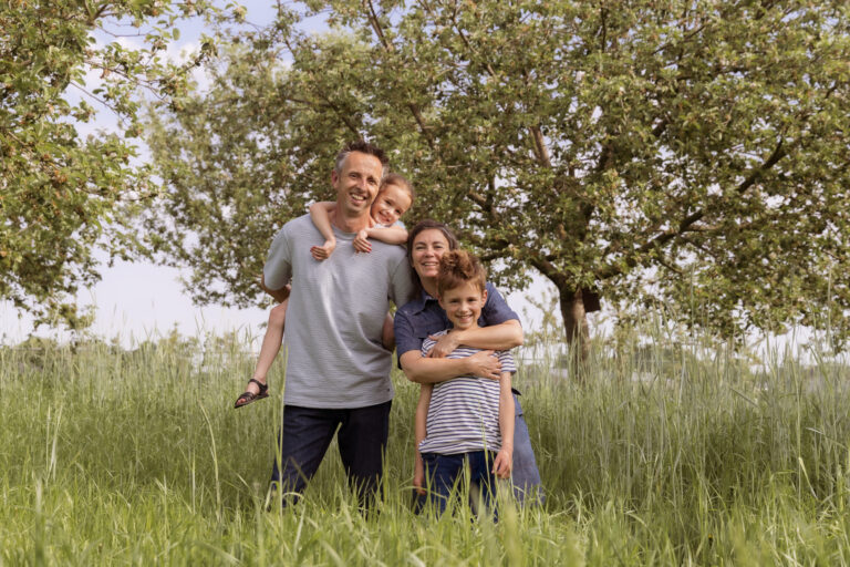 Gezin poseert lachend in grasveld met bomen op de achtergrond, zonnige dag.