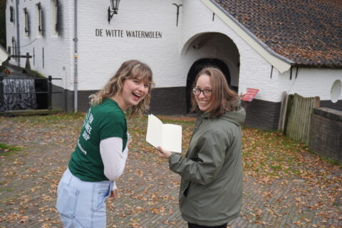 Twee lachende mensen bij De Witte Watermolen, één houdt een open notitieboek vast.