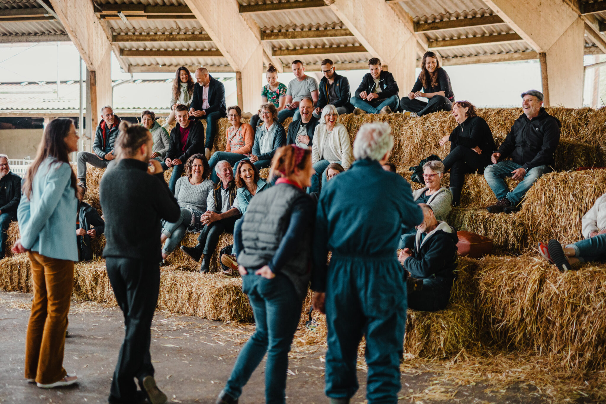 Groep mensen zit op strobalen in een schuur, luisterend naar sprekers.