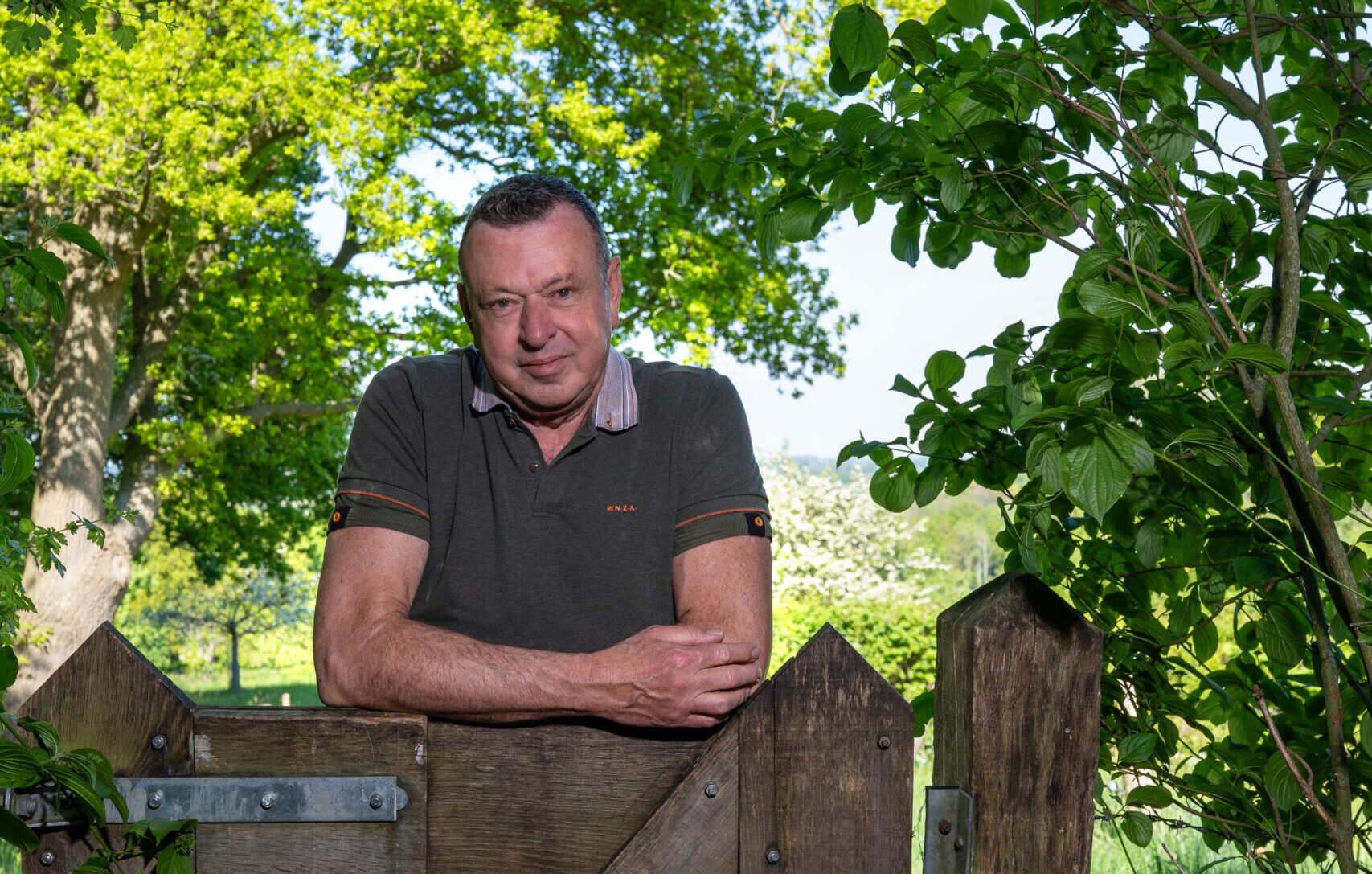 Man leunt op houten hek, omgeven door groene bomen en bladeren, zonnige dag.