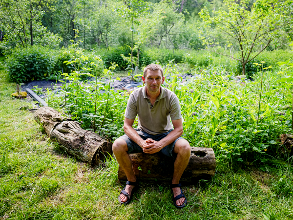 Man zit op een boomstam in groene tuinomgeving, omgeven door planten en bomen.